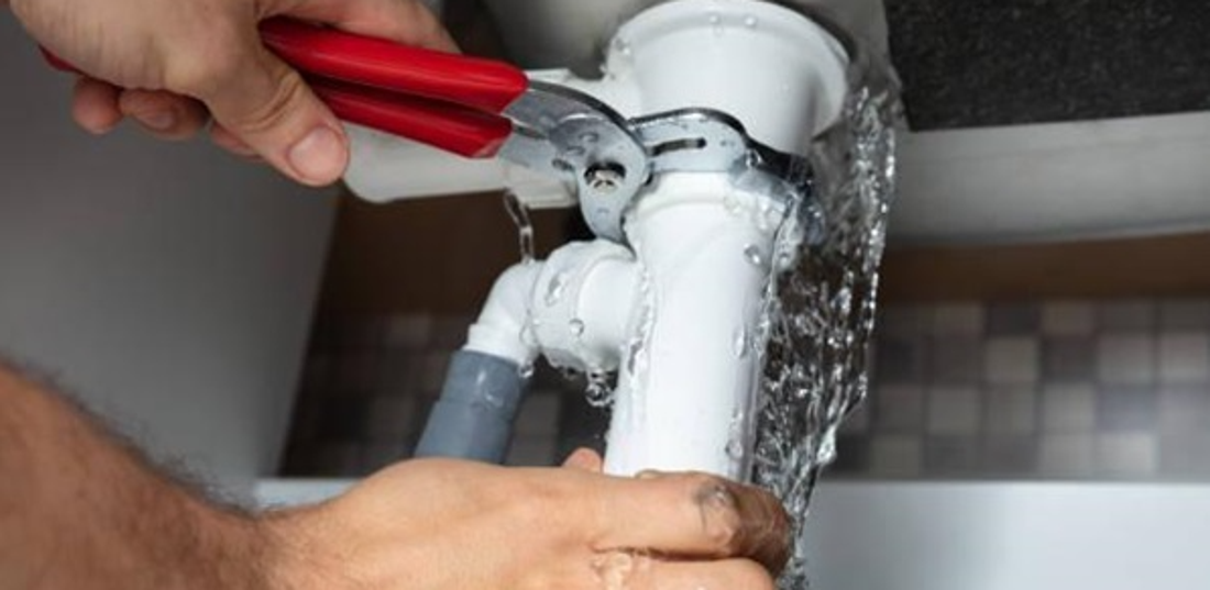 Plumber repairing an actively leaking pipe beneath a household sink.